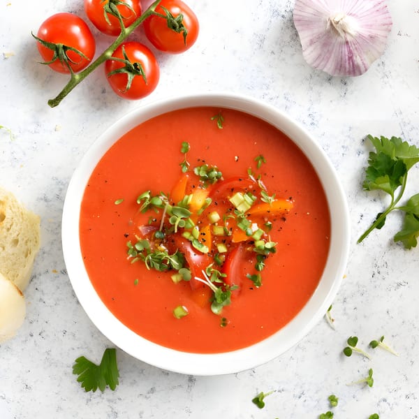 Vibrant red tomato soup in a white bowl, garnished with fresh herbs, alongside tomatoes and garlic.